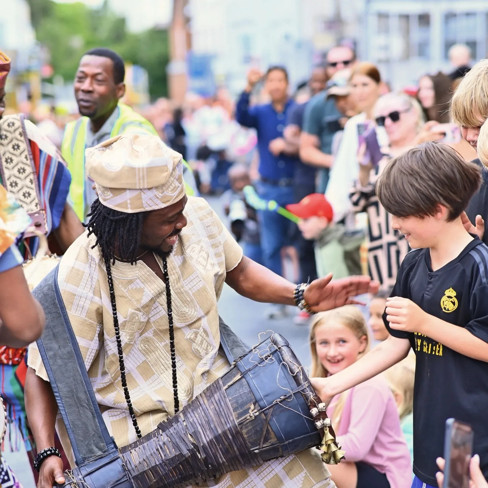 The founder lets a young boy play the talking drum at Colchester Carnival, as the crowd looks on