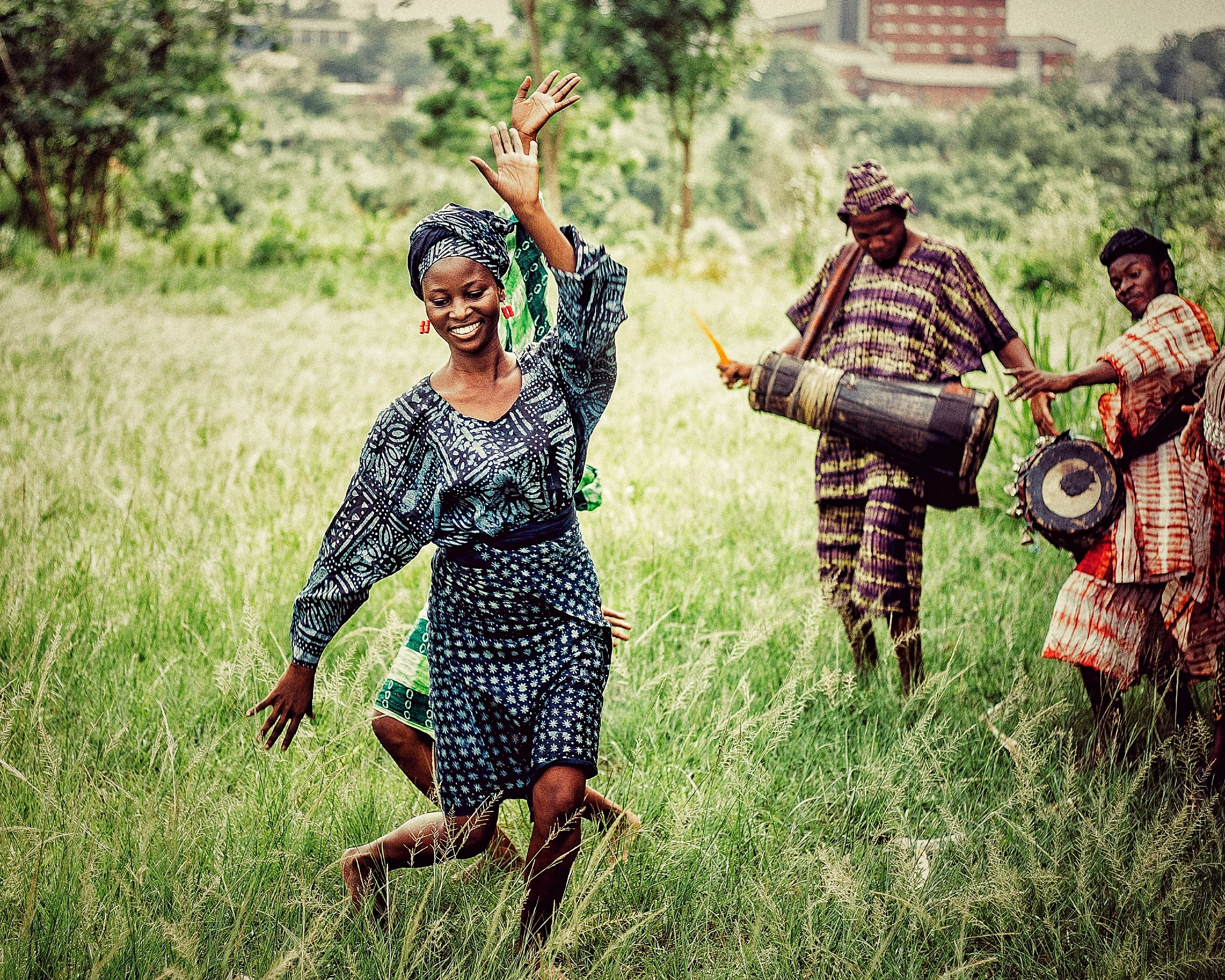 A dancer in àdìrẹ moves through tall grass while two drummers play the talking drum behind her